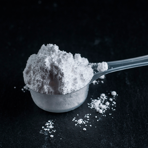 Colher medidora transparente com pó branco, representando suplemento alimentar em superfície escura.