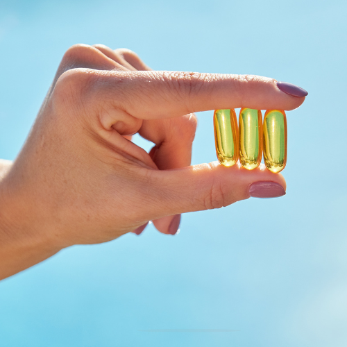Mão segurando três cápsulas amarelas de vitamina D entre os dedos, com céu azul claro ao fundo.