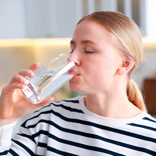 Mulher bebendo um copo de água em ambiente interno, com expressão tranquila e gesto de hidratação.