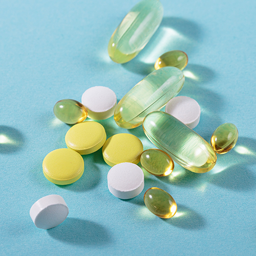 Comprimidos e cápsulas de diferentes tamanhos e cores sobre superfície azul, representando suplementos ou medicamentos.