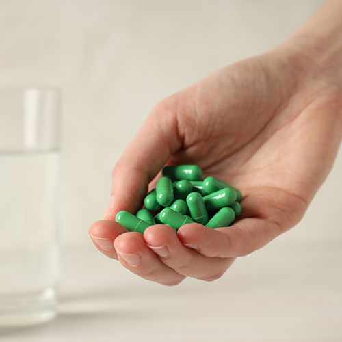 Mão segurando cápsulas verdes ao lado de frasco desfocado, representando uso de medicamentos ou diuréticos.
